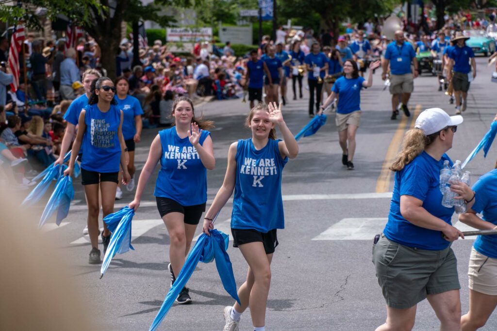 Several girls in blue Kennett High School t-shirts wave with blue banners in the Memorial Day Parade.