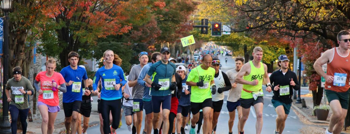 Men and women in athletic wear with numbers pinned to their shirts are running up Broad Street in Kennett Square in autumn